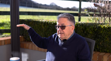 Man wearing sunglasses and a dark sweater sitting outdoors with greenery in the background
