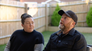 Two people sitting outdoors on a patio with a wooden fence and greenery in the background.