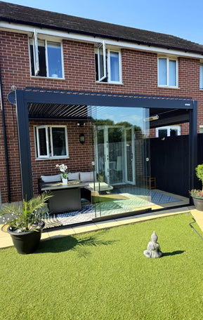 Modern patio area with glass wall, outdoor furniture, and plants in a residential setting. Black Pergola