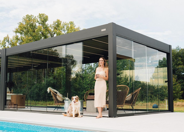 Woman standing inside a modern glass pergola with a dog by a pool, surrounded by trees.