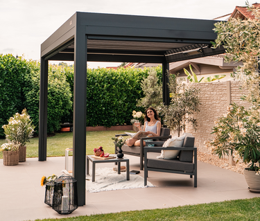 Woman sitting under a black pergola on a patio with outdoor furniture and plants.