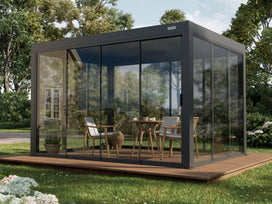PERGOLUX glass wall pergola with dark grey metal framing and clear tempered glass panels enclosing a wooden deck dining area