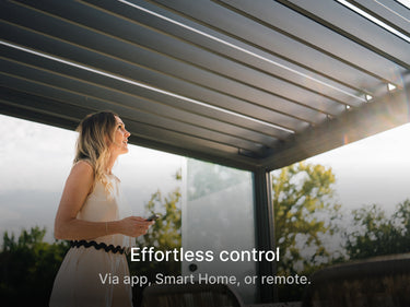 A medium-angle shot of a blonde woman in a white, sleeveless dress standing beneath a modern, dark louvered pergola. She is holding a small device, possibly a remote control or smartphone, and looking up toward the adjustable roof. Bright sunlight streams from the top right, creating lens flare. The background shows trees and a bright sky. A large text overlay at the bottom reads: "Effortless control" with a smaller line of text beneath it saying, "Via app, Smart Home, or remote."