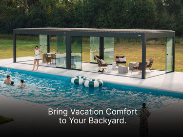 A wide, dark gray pergola with an adjustable louvered roof stands on a white deck adjacent to a rectangular swimming pool. The pergola has been enclosed with retractable glass walls, creating an elegant outdoor room.

Inside the enclosure, one section is set up as a dining area with a table and chairs, and the other is a lounge area with modern wicker chairs and small side tables.

Outside, people are swimming and playing in the pool, with two large green and white striped inflatable rings floating nearby. 
