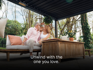 A middle-aged couple is sitting closely together on a white outdoor sofa with a coral-colored pillow, beneath a large, dark louvered pergola. The man in a pink shirt is looking affectionately at the woman in a white tank top. They are next to a wooden outdoor coffee or dining table that holds a platter of food and a potted plant. The setting is a lush backyard with mature trees and greenery visible in the background. A text overlay at the bottom of the image reads: "Unwind with the one you love."
