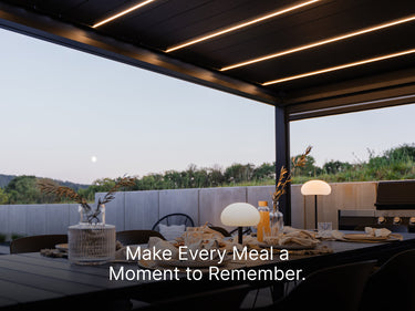 A close-up view of an elegantly set outdoor dining table beneath a dark-colored pergola with bright, integrated LED strip lighting. The table, which appears to be dark gray or black, is set for a meal with glassware, cloth napkins, and decorative vases containing dried grass or wheat. Two modern white table lamps provide soft, warm light. In the background, there is a low concrete wall and a natural landscape with rolling hills and trees visible under a twilight sky with a visible full moon. A text overlay 