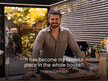 A man stands and smiles in a modern outdoor kitchen beneath a stylish pergola, with food and drinks on the counter. Text reads: "It has become my favorite place in the whole house." Thore Schölermann.