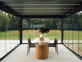 Modern indoor-outdoor space with a dining table and chairs near large glass windows under a pergolux pergola.