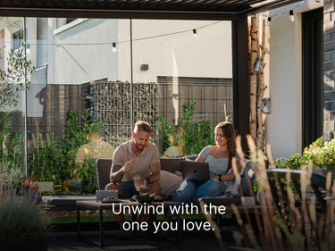 
A smiling young couple is relaxing together on a dark gray outdoor sofa beneath a covered patio or pergola. The man is holding a drink and looking at the woman, who is using a laptop. They are seated next to a small black coffee table that holds a tablet and snacks. The space is partially enclosed by large glass panels and decorated with potted plants, string lights, and natural-looking wooden columns. The background shows a contemporary building and a gabion wall. A text overlay at the bottom reads: "Unwi