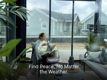 An image taken from the cozy interior of a glass-enclosed pergola or sunroom on a rainy day. A young woman is seated comfortably in a chair, wrapped in a blanket, wearing a white sweater, reading a book, and sipping from a mug. The glass walls are streaked with heavy rain, obscuring the view of the suburban houses outside, yet the interior remains bright and dry. The space is furnished with modern outdoor furniture and a coffee table. The image has a logo overlay for "RainLUX™" and a text overlay at the bot