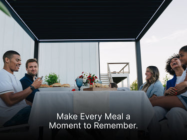 
A diverse group of five friends or family members are gathered around a dining table for a meal beneath a dark, modern pergola. The table is covered with a white tablecloth and set with plates of food (burgers or sandwiches), a centerpiece of red and white flowers, and glassware. The people are laughing and smiling, enjoying each other's company. The setting is a backyard patio next to a white house, with a wooden deck and stairs visible through the pergola's frame. A text overlay at the bottom reads: "Mak