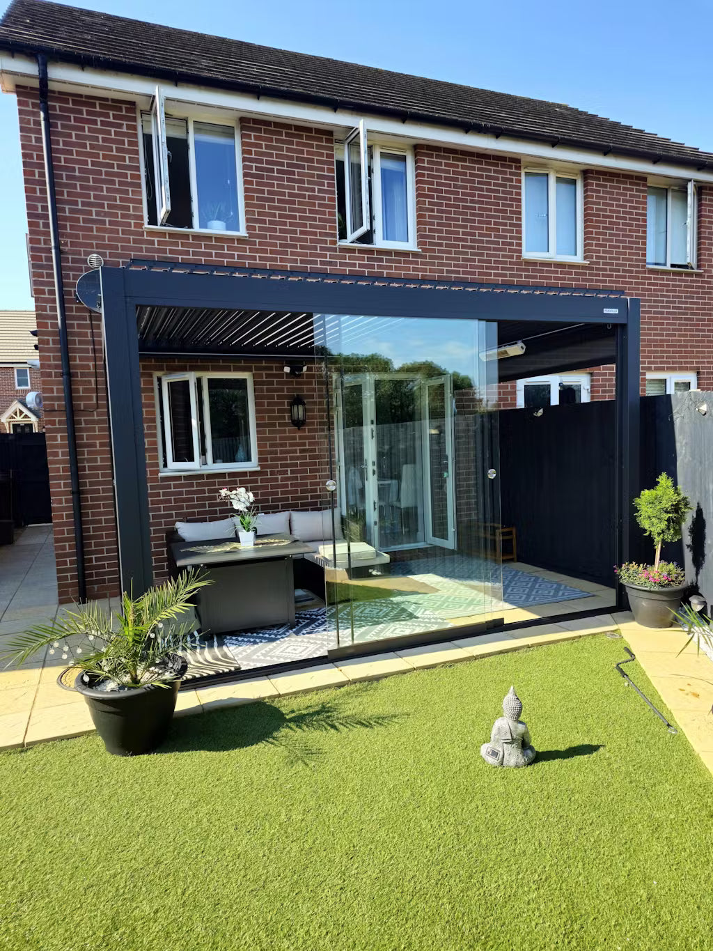 Modern patio area with glass wall, outdoor furniture, and plants in a residential setting. Black Pergola